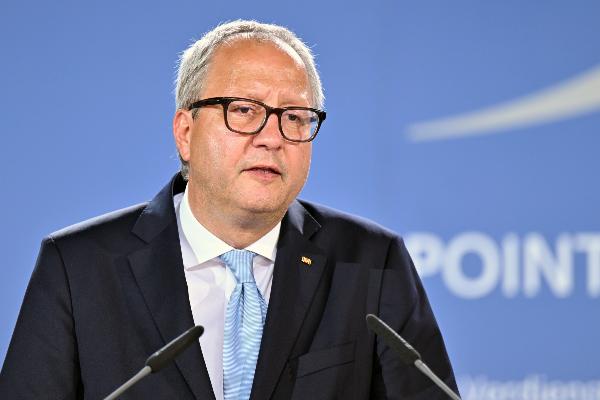 Ein Mann in Anzug und Krawatte spricht bei einer Pressekonferenz vor blauem Hintergrund mit Mikrofonen.
