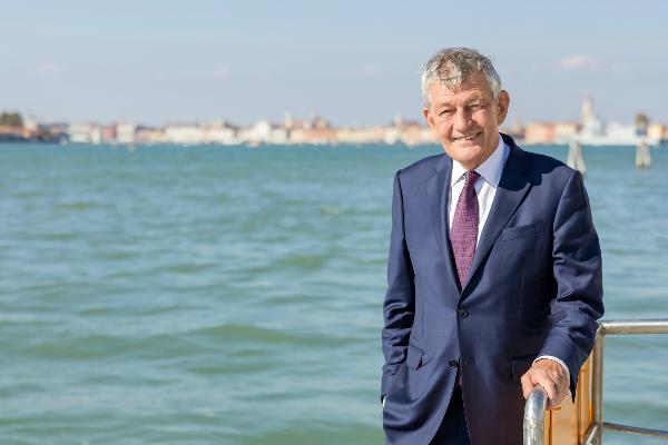 Professor Huber is standing in the sunlight at a railing in front of a body of water. He is wearing a dark blue suit. In the background, you can see some sailboats and buildings in Venice.