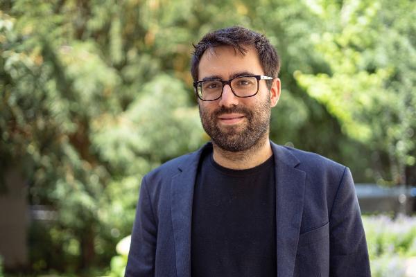 Professional portrait of Professor Sebastian Bender wearing glasses, a dark blazer, and a black T-shirt in a lush green park and garden setting.