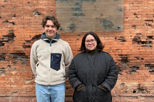 Simon Theodor Fetscher and Victoria Lobato stand in front of the Salinengebäude of the University of Munich, a brick wall featuring a glass relief with the text WUNDEN DER ERINNERUNG (Wounds of Memory). The wall shows signs of peeling and damage.