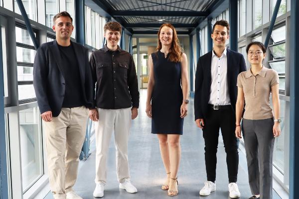Five members of a team stand smiling in a modern glass corridor of an office and research building. The professional group photo highlights collaboration, innovation, diversity, and a welcoming company culture in a contemporary workplace.