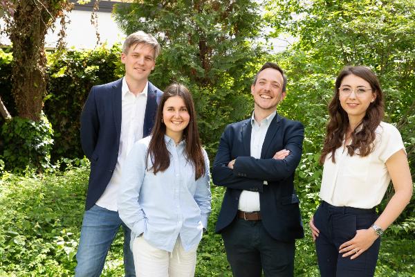 Four smiling people, two men and two women, stand in formal dress in a green garden with trees in the background.