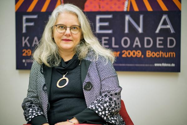 Porträt der Theaterwissenschaftlerin Professor Meike Wagner. Sie sitzt vor einem großen Figurentheaterposter. Sie trägt eine Brille, einen schwarzen Pullover und eine graumeliert Jacke.
