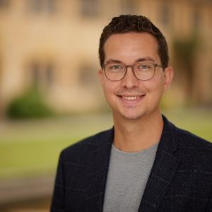 Ein lächelnder Mann mit Brille und dunklem Haar trägt ein dunkelblaues Sakko über einem grauen Pullover. Unscharfer Hintergrund mit grüner Natur und historischer Architektur.