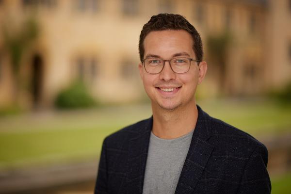 Ein lächelnder Mann mit Brille und dunklem Haar trägt ein dunkelblaues Sakko über einem grauen Pullover. Unscharfer Hintergrund mit grüner Natur und historischer Architektur.