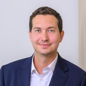 Porträt eines lächelnden Mannes im blauen Anzug vor heller Wand, geeignet für Business- oder Teamprofil.