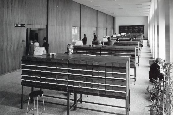 Schwarzweißaufnahme eines Bibliotheksraums der 1960er-Jahre mit Karteikästen, Lesetischen und mehreren Personen bei der Recherche.