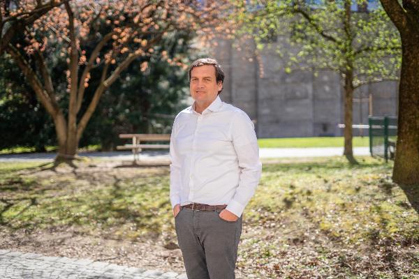 Ein Mann in weißem Hemd steht mit Händen in den Hosentaschen in einem sonnigen Park mit Bäumen im Hintergrund.